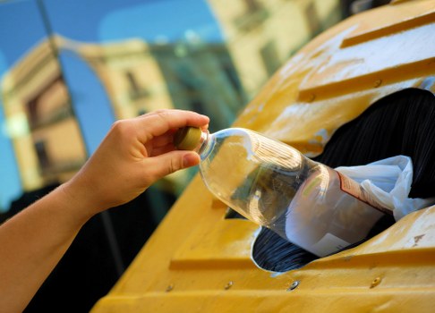 A close-up view of a person's hand placing a transparent glass bottle into a dedicated recycling or waste container on a yellow-painted surface, likely part of a waste disposal area outside a building. The bottle appears to be a standard plastic beverage container with a yellowish cap, and the surface on which it rests shows signs of weathering. In the background, blurred images of a building façade and greenery can be seen, indicating an outdoor setting. The lighting suggests natural daylight, highlighting the textures of the container and the surface below. This image relates to rubbish removal services in Downham, such as those offered by Commercial Waste Downham, emphasizing the collection and disposal of recyclable bottles and other waste materials in a residential or commercial context.