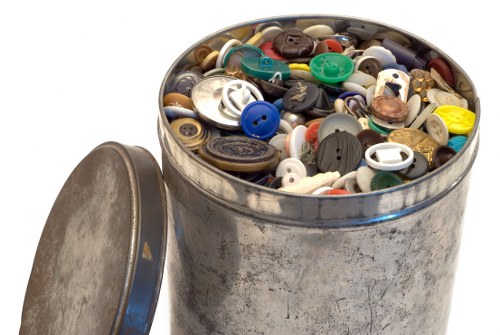A round, metal container with a slightly weathered, tarnished surface, filled with a large assortment of assorted buttons in various sizes, shapes, and colors, including black, white, blue, red, green, and beige. Some buttons are plastic, with smooth or textured finishes, and a few feature metallic or decorative elements. The container is positioned outdoors on a surface, with its matching metal lid resting on the left side, partly leaning against the container. The background is plain white, emphasizing the focus on the buttons and the metallic container, which suggests it may be used for collection or disposal of small household items in a rubbish removal context, such as cleaning out storage or a workshop.