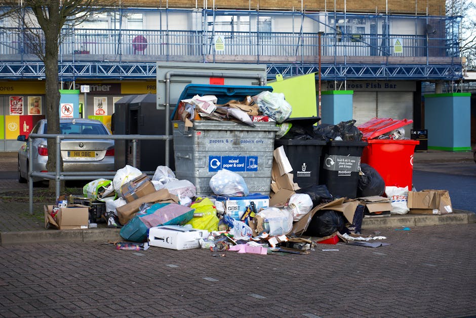 A street scene showing a worker in a high-visibility yellow and orange jacket operating a large, red waste collection vehicle, which is positioned on the roadside with its rear open for rubbish loadin