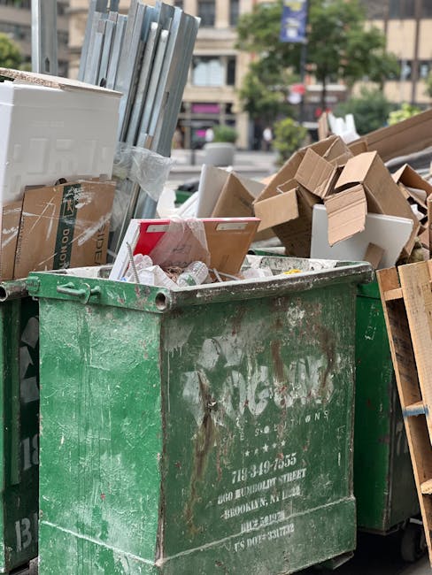A large, weathered red metal skip positioned in a narrow urban alleyway, situated against a dark green wall on the left and a light grey concrete wall on the right. The skip shows signs of rust and pa