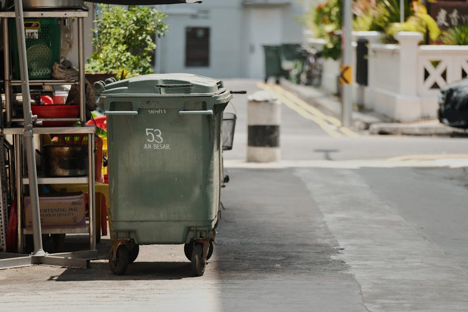 A black wheeled commercial waste container placed on the sidewalk in front of a storefront restaurant with a red-brown facade and large windows. The container, marked with white lettering indicating '