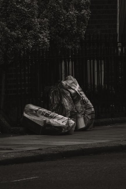 A collection of black rubbish bags, some torn open, stacked and scattered in front of a red metal door affixed to a textured, off-white exterior wall. The bags appear to contain various household wast